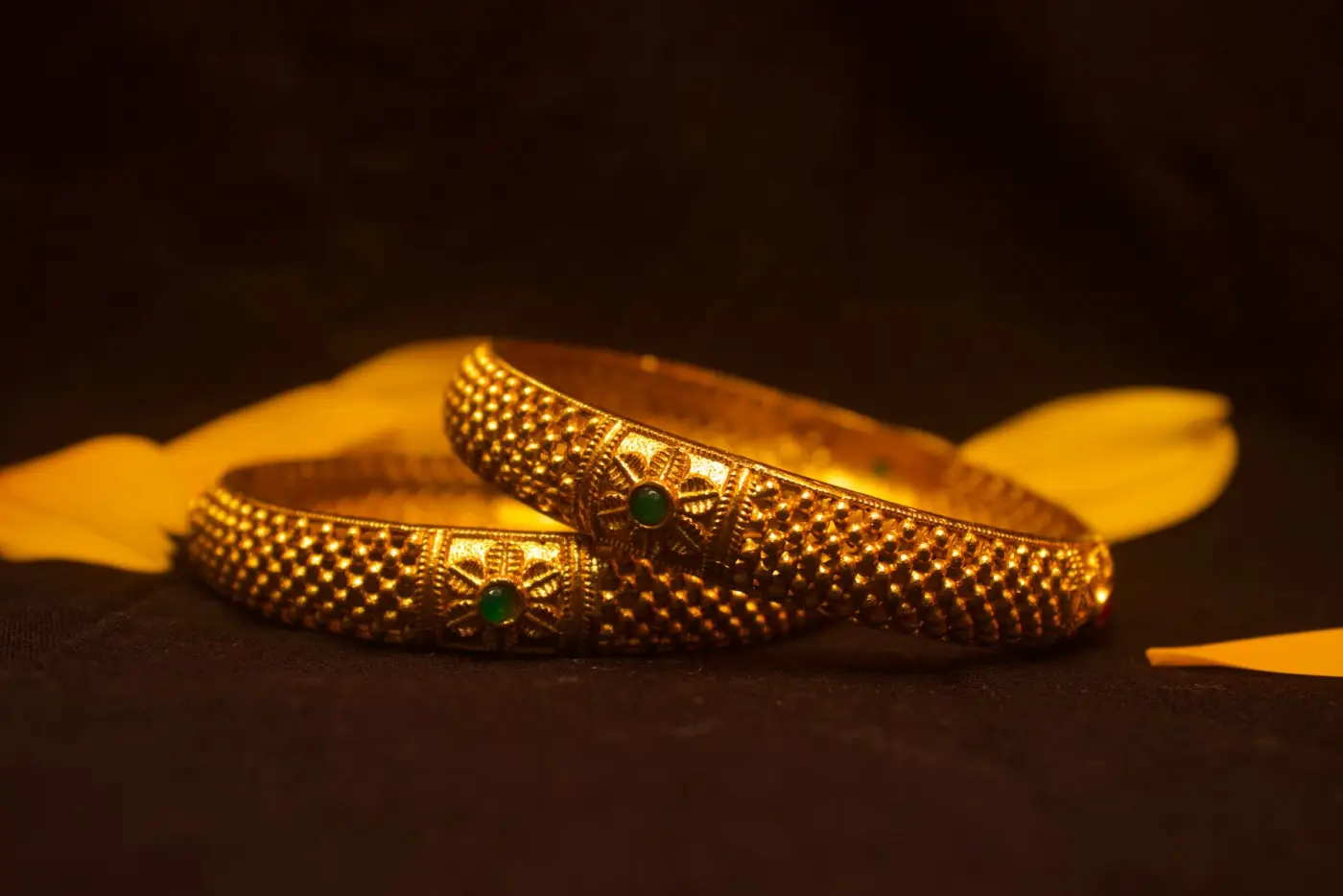 Two gold bangles with fine filigree and small green stones, on a dark cloth